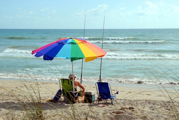 An elderly man sits under a rainbow umbrella fishing on the beach